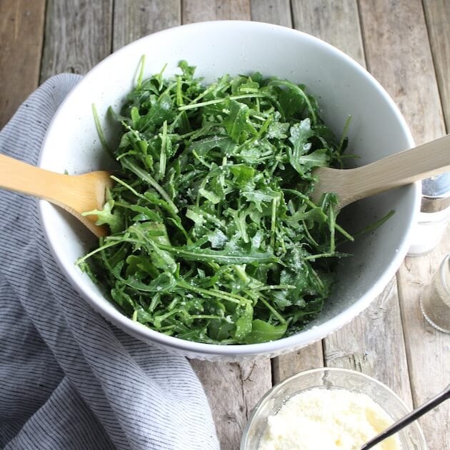 Arugula Salad With Parmesan &amp; Lemon in white serving bowl with wooden spoons