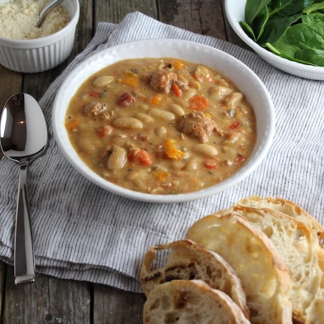 Bowl of bean soup on a table with fresh spinach, parmesan cheese, and french bread