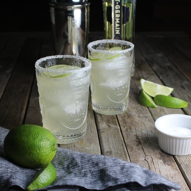 Two Elderflower Margaritas on farm table with limes and salt on table