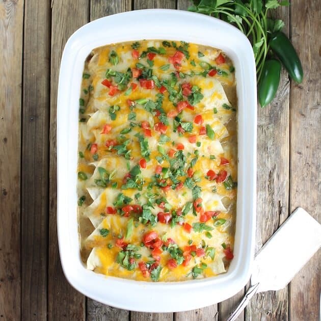 Casserole dish full of Cheesy Chicken Enchiladas with spatula and jalapenos