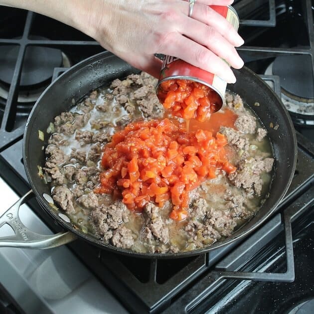 Adding canned tomatoes to browned lamb meat in saute pan