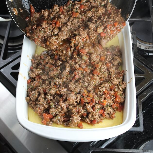 Adding lamb lasagna meat mixture to casserole dish with layer of polenta in the bottom