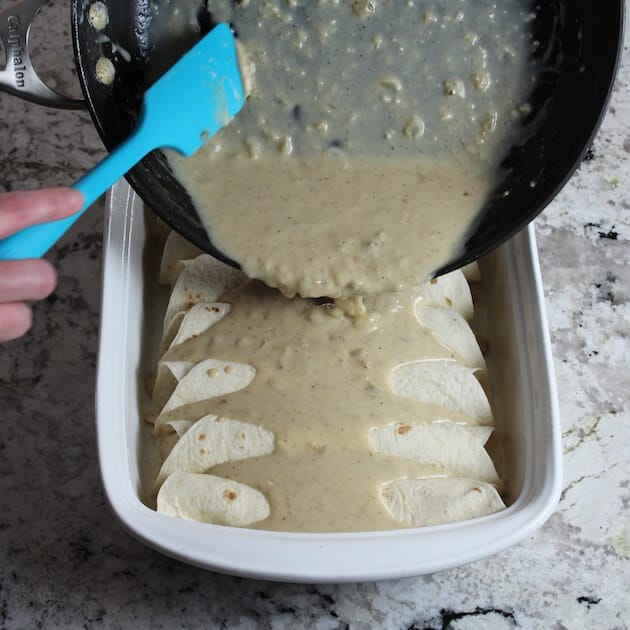 Pouring cheesy sauce on top of tortillas in casserole dish for enchiladas