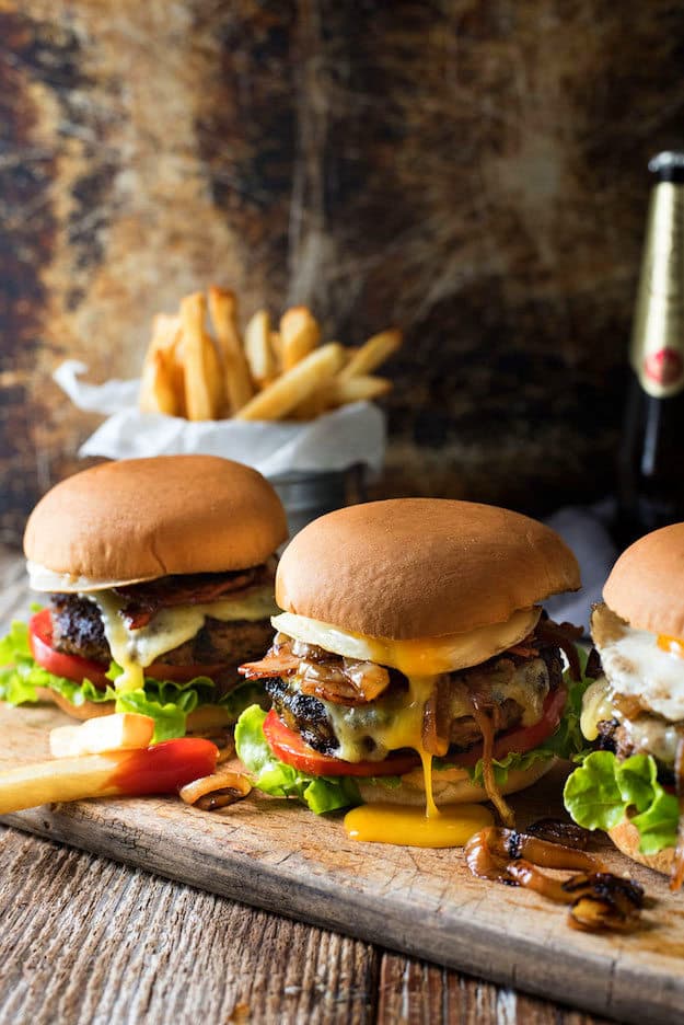 Three cheesy burgers lined up on a cutting board with french fries in background