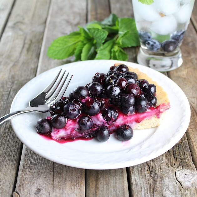 Slice of Mascarpone Blueberry Tart on plate with glass of water and mint