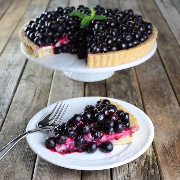 Piece of Blueberry Tart on plate with whole tart on cake platter in background 