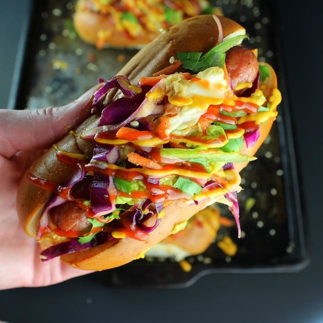Hand holding a Korean Slaw Hot Dog with bright colored Asian slaw, mustard, and ketchup