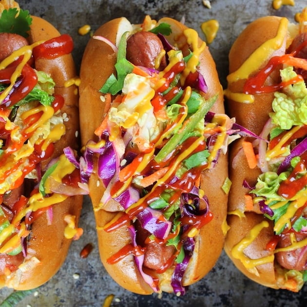 Three Korean Slaw Hot Dogs on cookie sheet with bright colored slaw, mustard, and ketchup