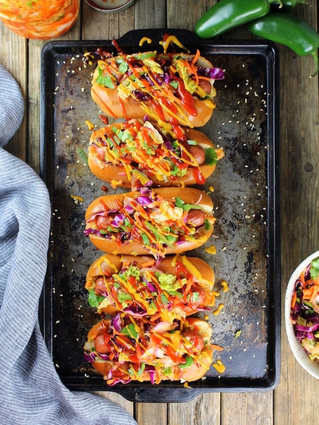 Five Korean Slaw Hot Dogs on cookie sheet with bright colored slaw, mustard, and ketchup