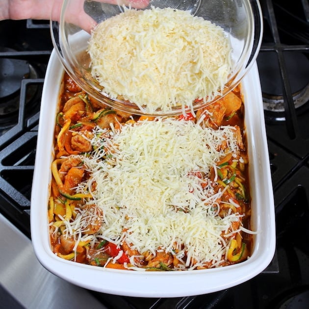 Adding shredded cheddar to the top of a casserole dish with italian sausage, peppers, and zoodles.