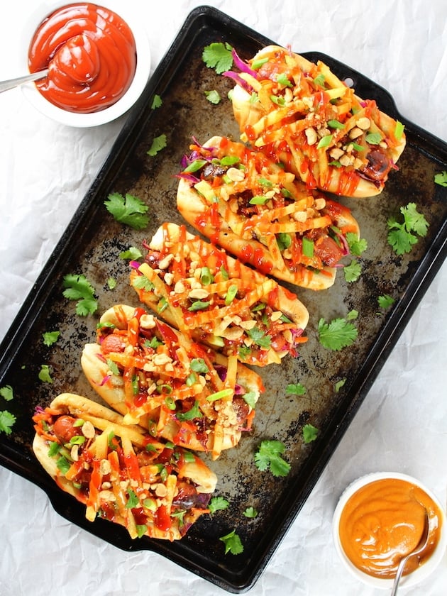 5 asian hot dogs lined up on a baking sheet, with slaw, french fries, and spicy ketchup