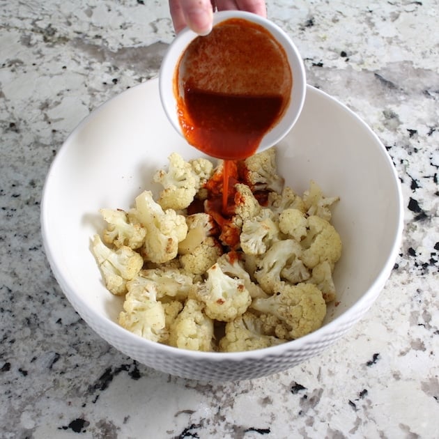 Pouring buffalo sauce over bowlful of roasted cauliflower florets