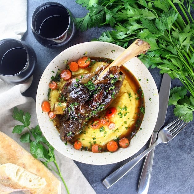 Lamb shank meal with bread, silverware, red wine, and parsley garnish