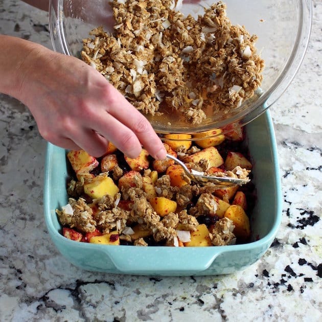 Adding crumble mixture to mangoes and blueberries in a baking dish