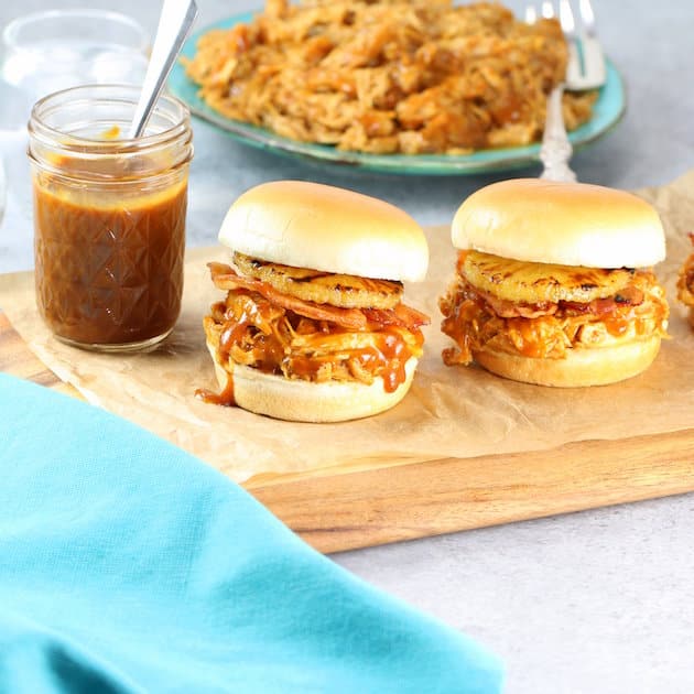 Two Hawaiian chicken and bacon sliders on cutting board with mason jar of BBQ sauce