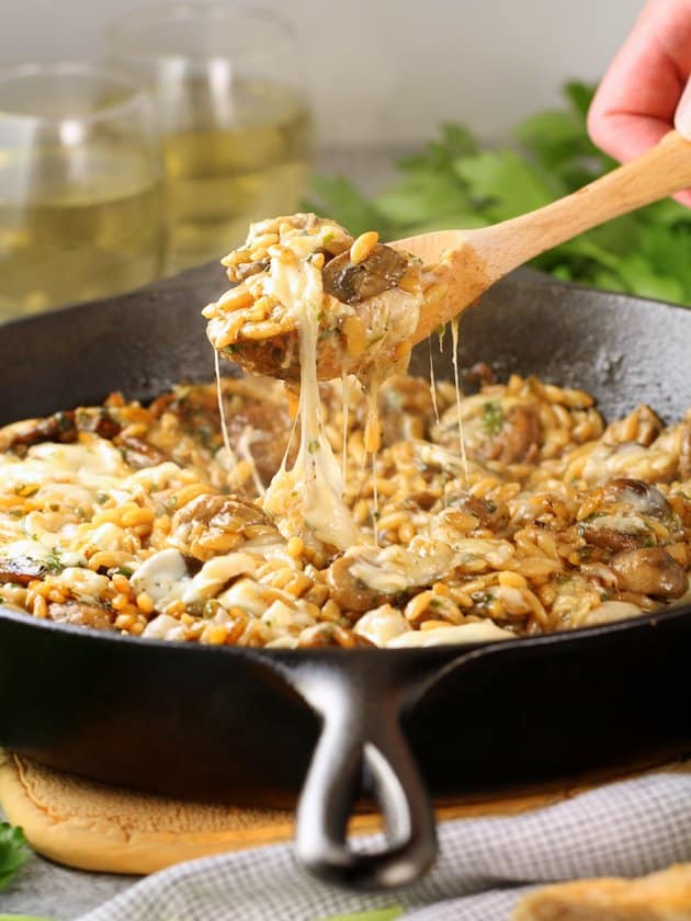 Wooden spoon lifting cheesy scoop of Marsala Chicken Orzo out of a cast iron Skillet