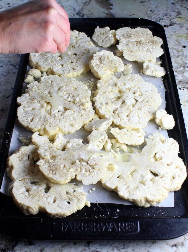 Adding salt and pepper to sliced cauliflower steaks on baking sheet