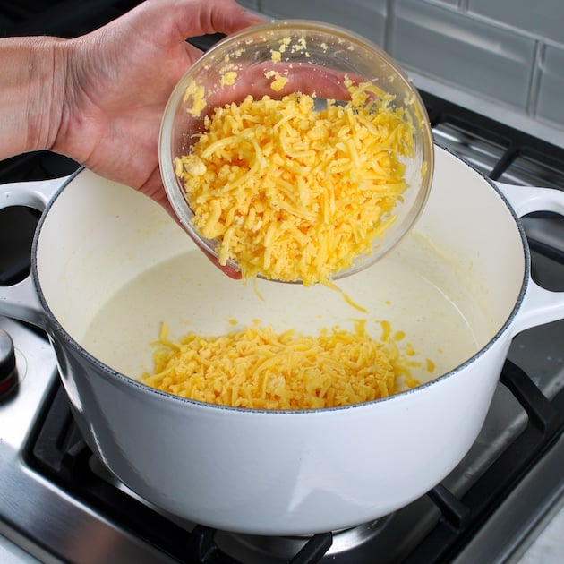 Adding shredded cheddar to a large white soup pot with warmed milk mixture