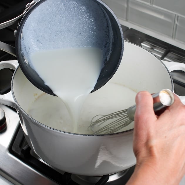 Pouring warm milk into large white soup pot while whisking, for mac and cheese
