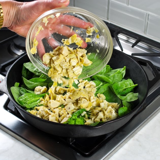 adding artichokes to a cast iron skillet with fresh baby spinach