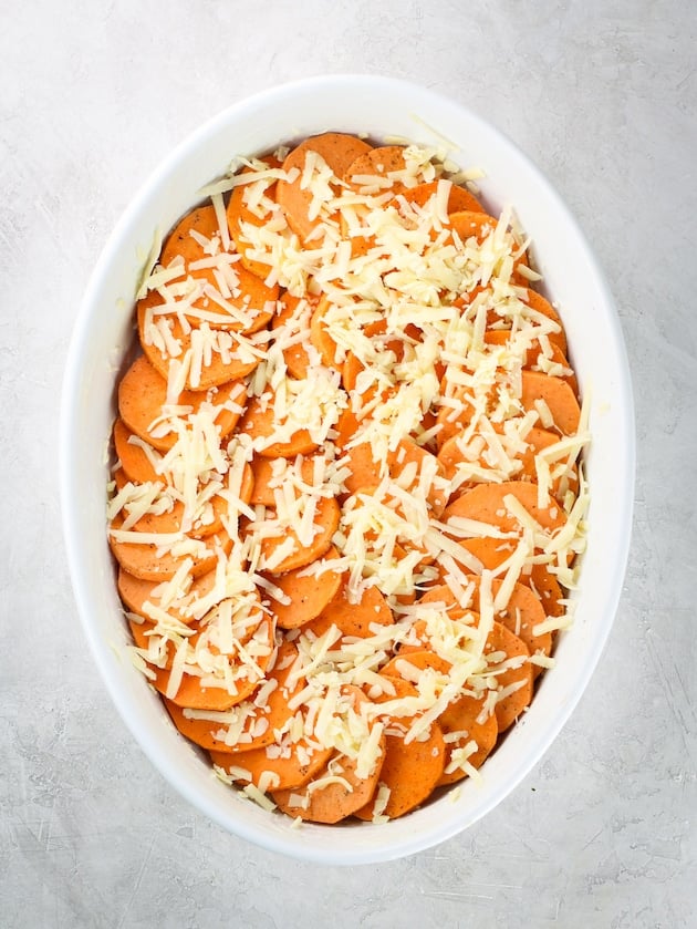 Sliced sweet potatoes arranged in a gratin baking dish with shredded Gruyere cheese.