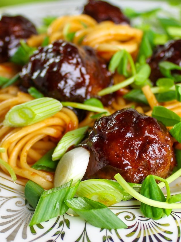 Very close photo of a fancy china plate with Asian noodles, meatballs, pea shoots, and green onions