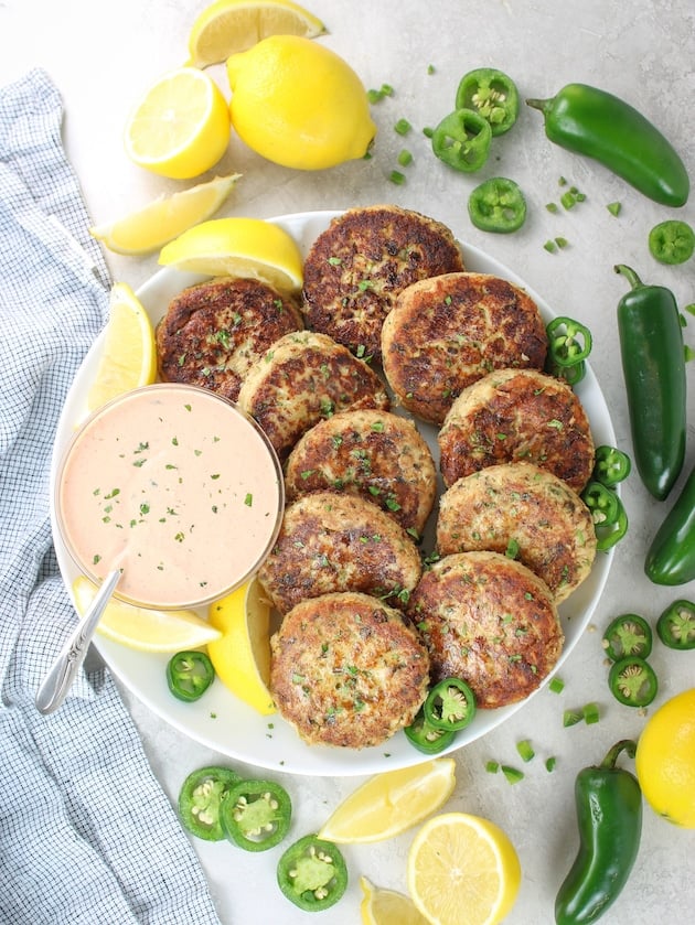 A wide shot of 10 Tuna Patties arranged on a plate with Sririacha aioli sauce in a bowl next to them and cut up lemons, whole lemons, slice jalapeños and whole jalapeños.