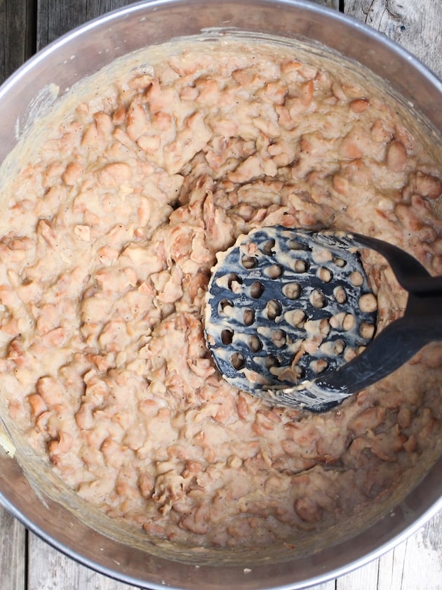 Cooked beans being smashed with a potato smasher.