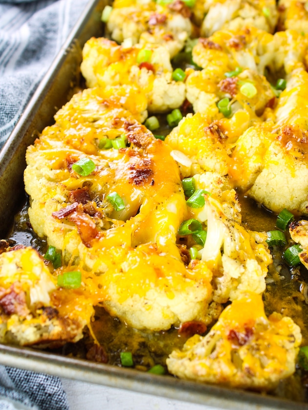 A lose up photo of Cauliflower steaks roasted on a baking sheet and topped with cheddar cheese, bacon and green onions.