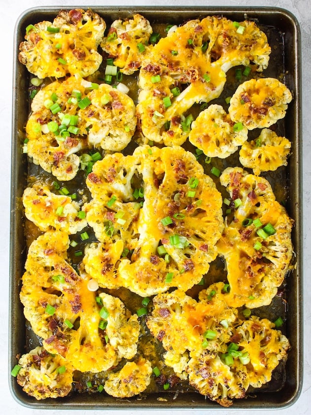 A sheet pan with roasted cauliflower steaks topped with cheddar cheese, bacon and green onions.