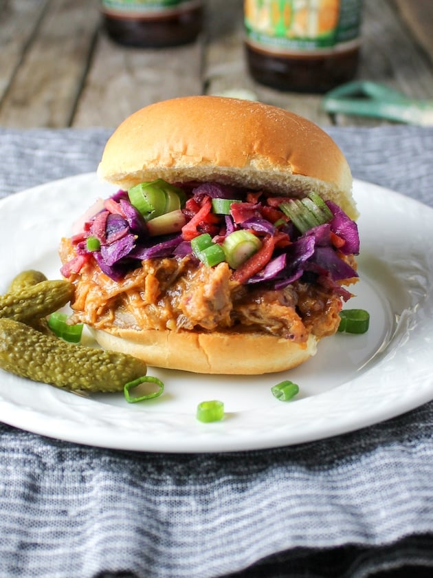 A plate with a pulled pork sandwich and a bottle of beer in the background.