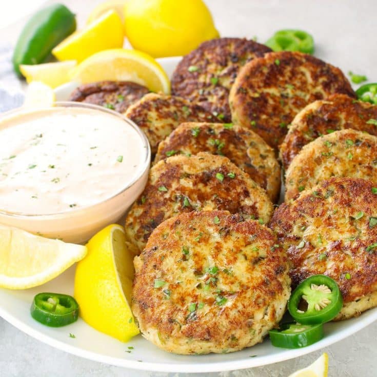 10 pan-fried tuna cakes on a platter with a creamy sauce and lemon wedges.