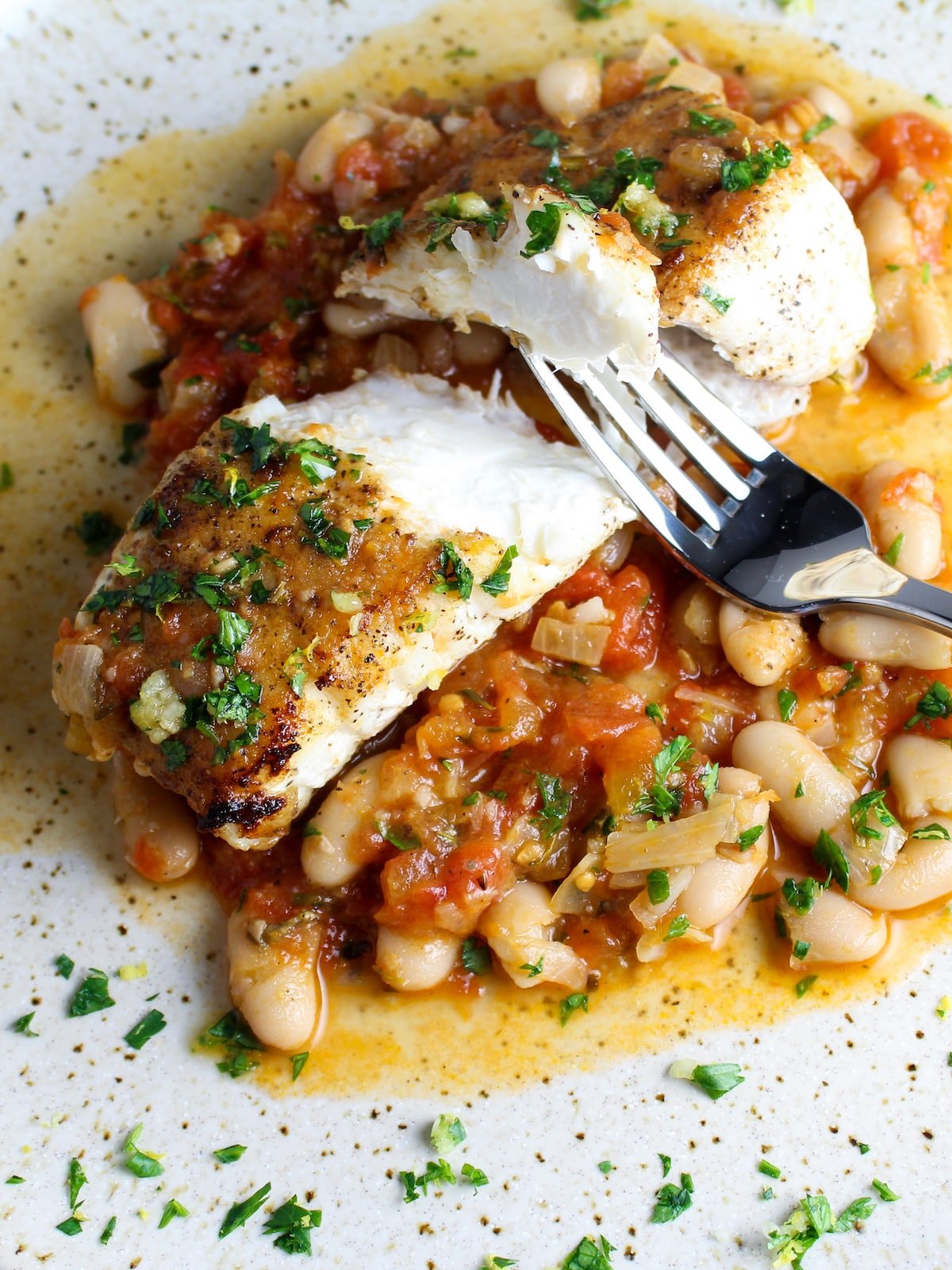 One piece of cooked flaky white fish cut in half on a plate with a fork over sautéed cannellini beans.