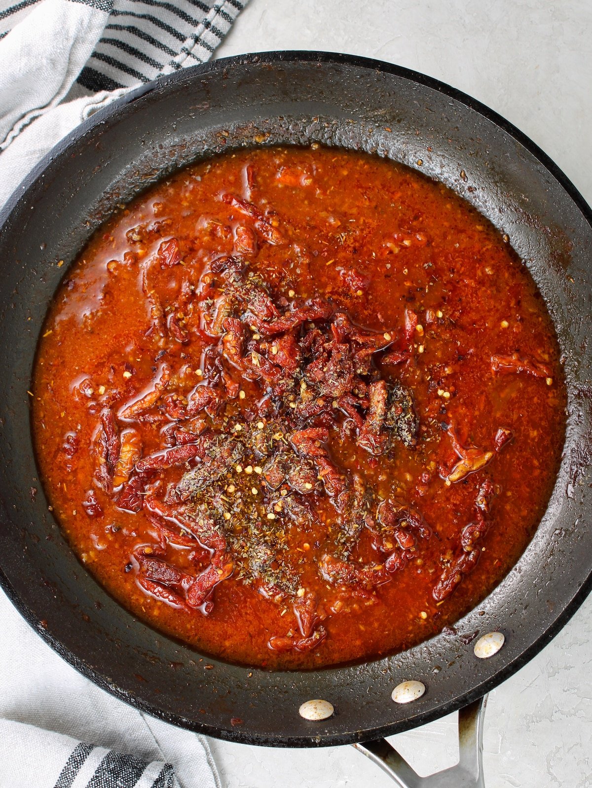 A saute pan with chicken stock, sun-dried tomatoes, spices, and herbs.