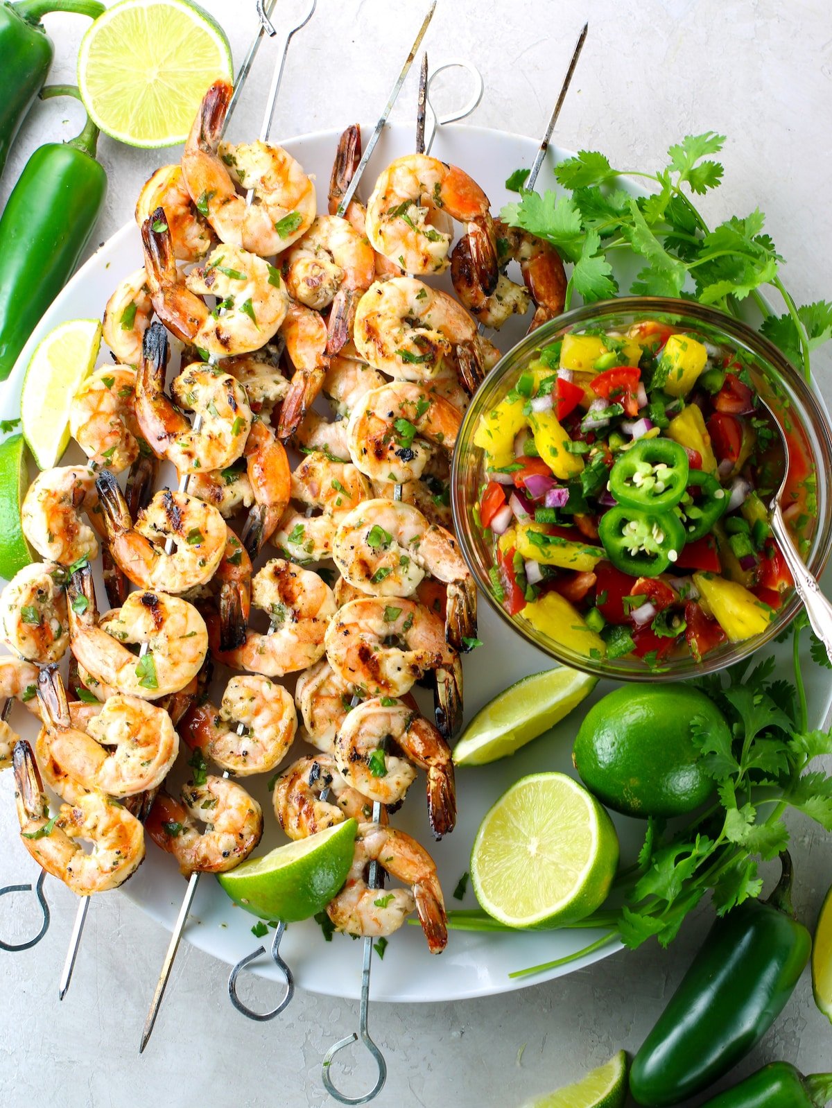 A platter of grilled shrimp on skewers with pineapple salsa and lime, cilantro, and jalapeno garnish.