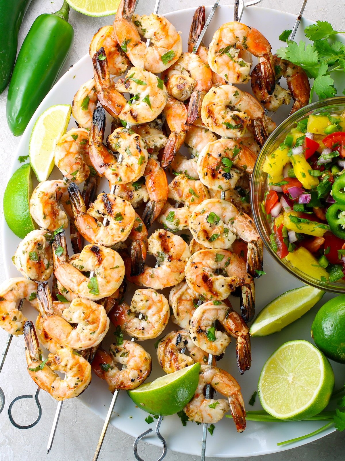 A platter of grilled shrimp on skewers with pineapple salsa and lime, cilantro, and jalapeno garnish.