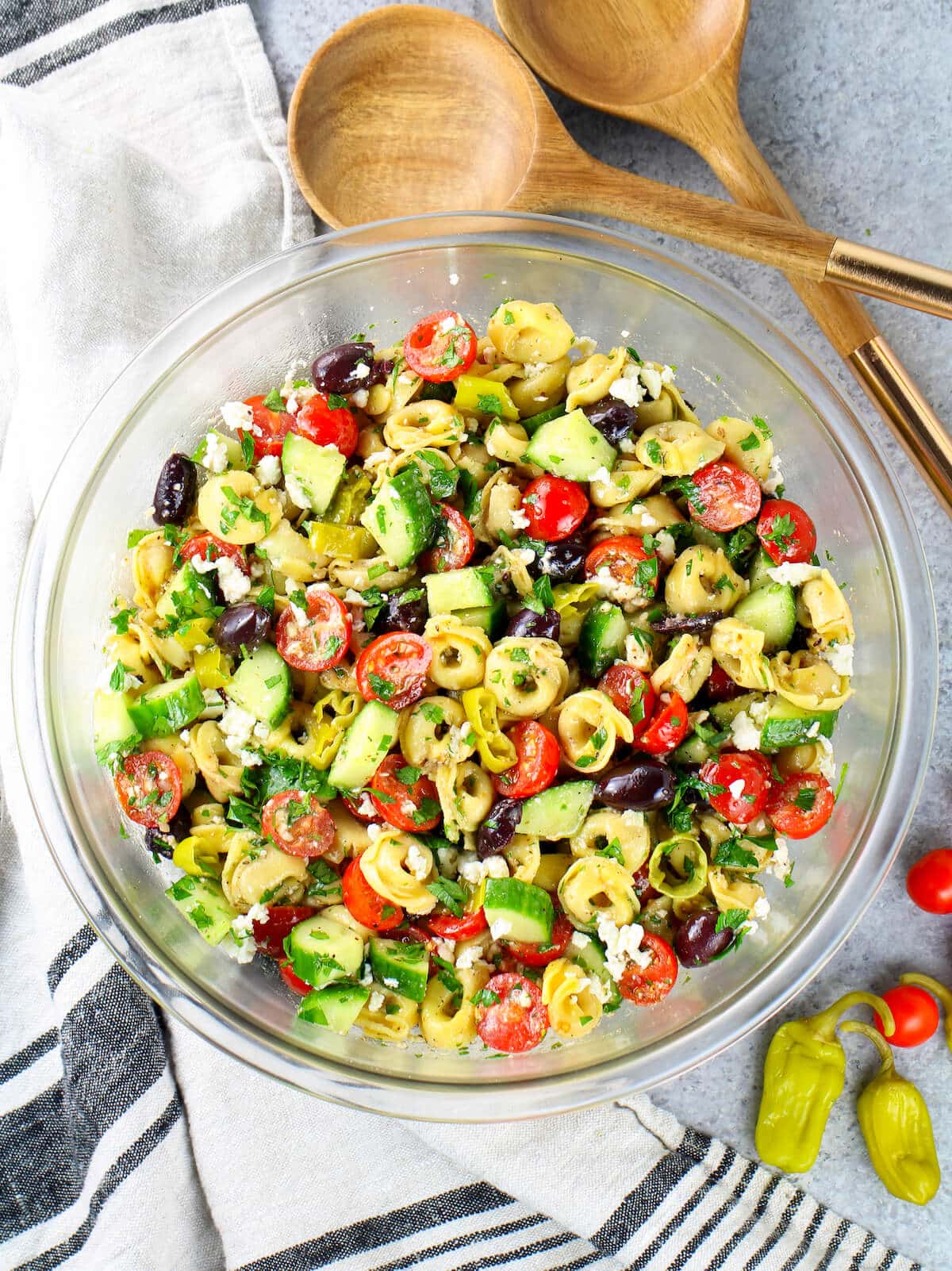 A large glass bowl with pasta, veggies, and dressing mixed together with serving utensils on the side.