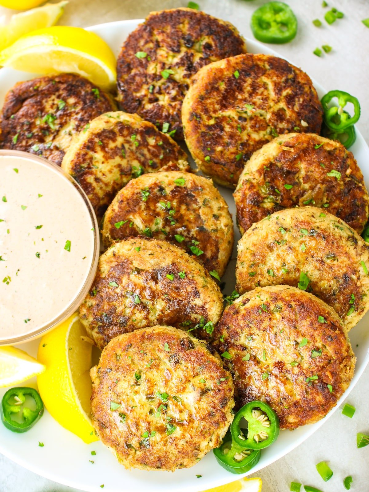10 tuna cakes arranged on a plate with Sriracha aioli sauce in a bowl next to them.