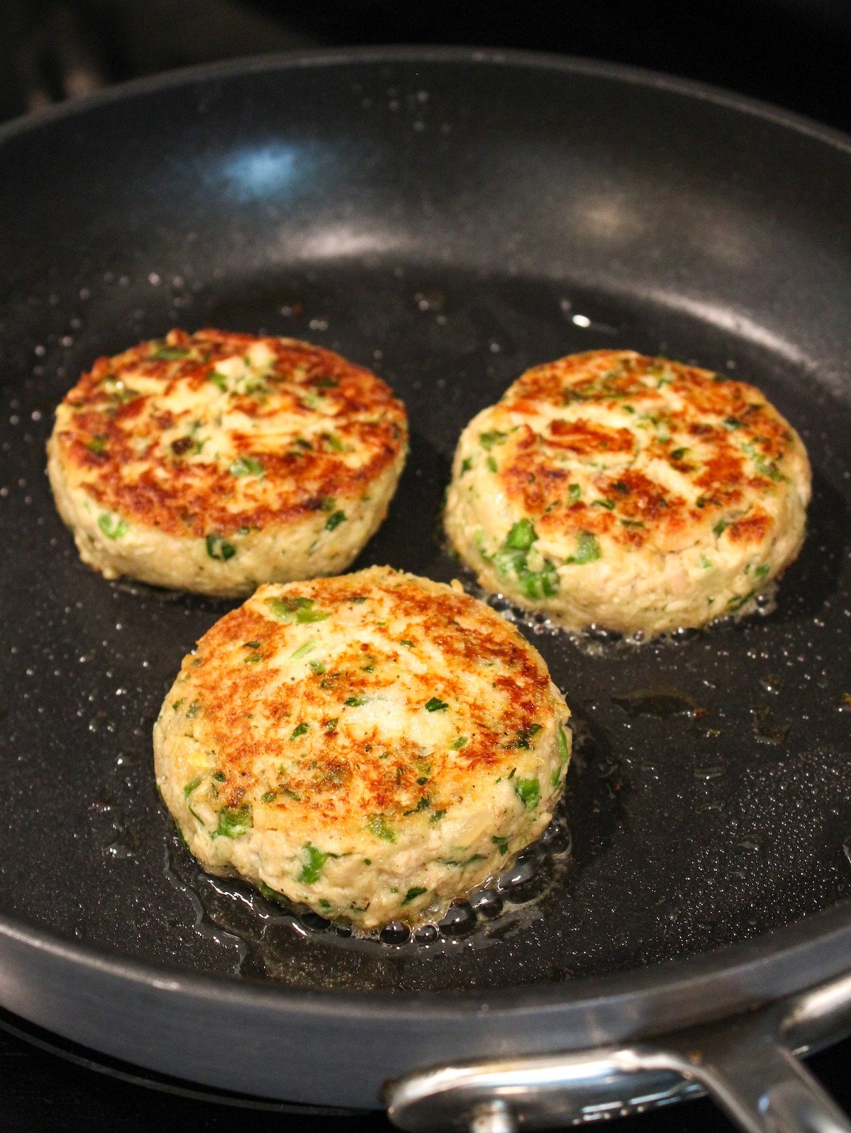 Three tuna patties frying in a nonstick skillet on the stove top.