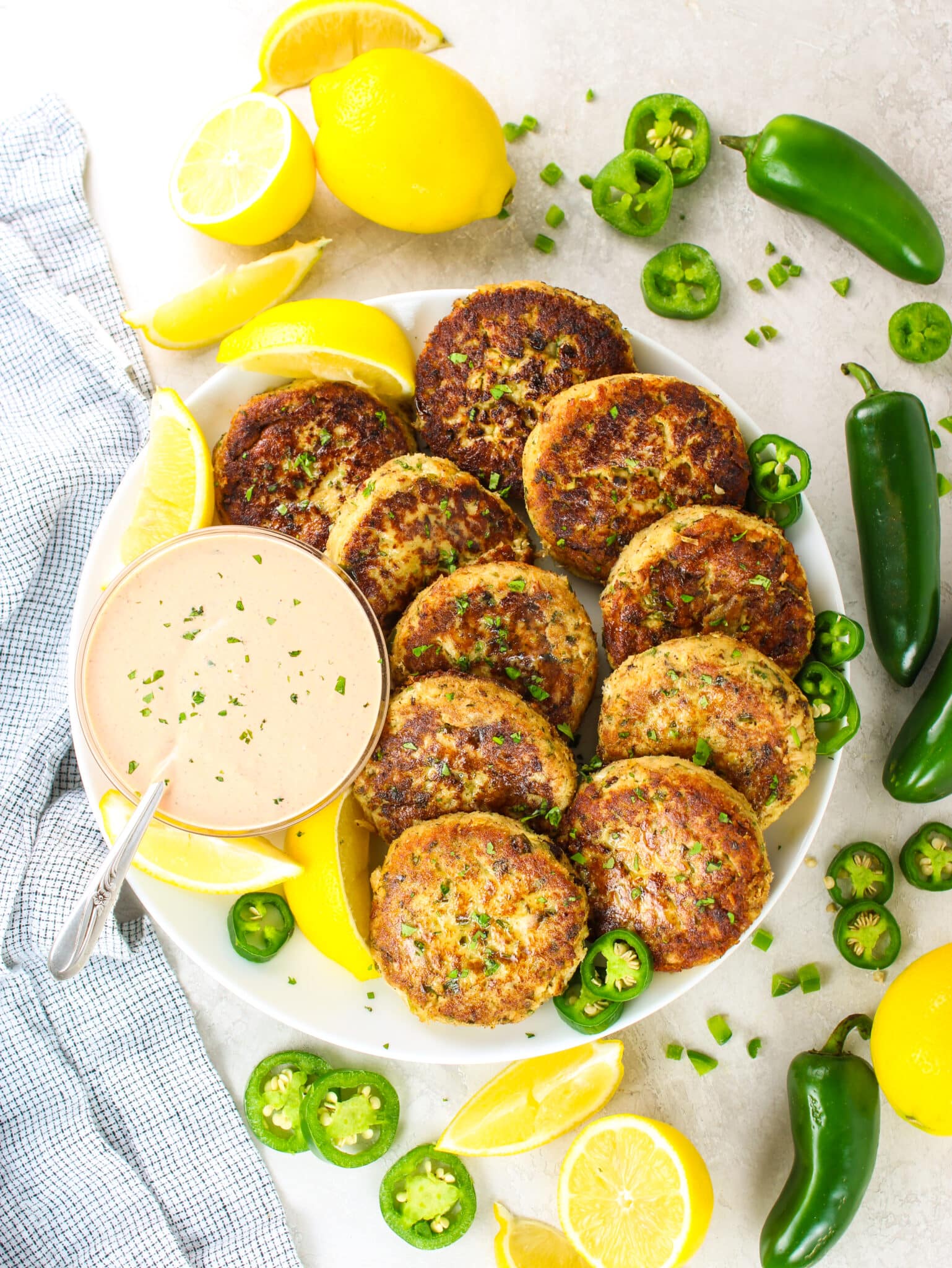 A wide shot of 10 tuna cakes arranged on a plate with Sriracha aioli sauce in a bowl next to them and cut up lemons, whole lemons, slice jalapeños and whole jalapeños.