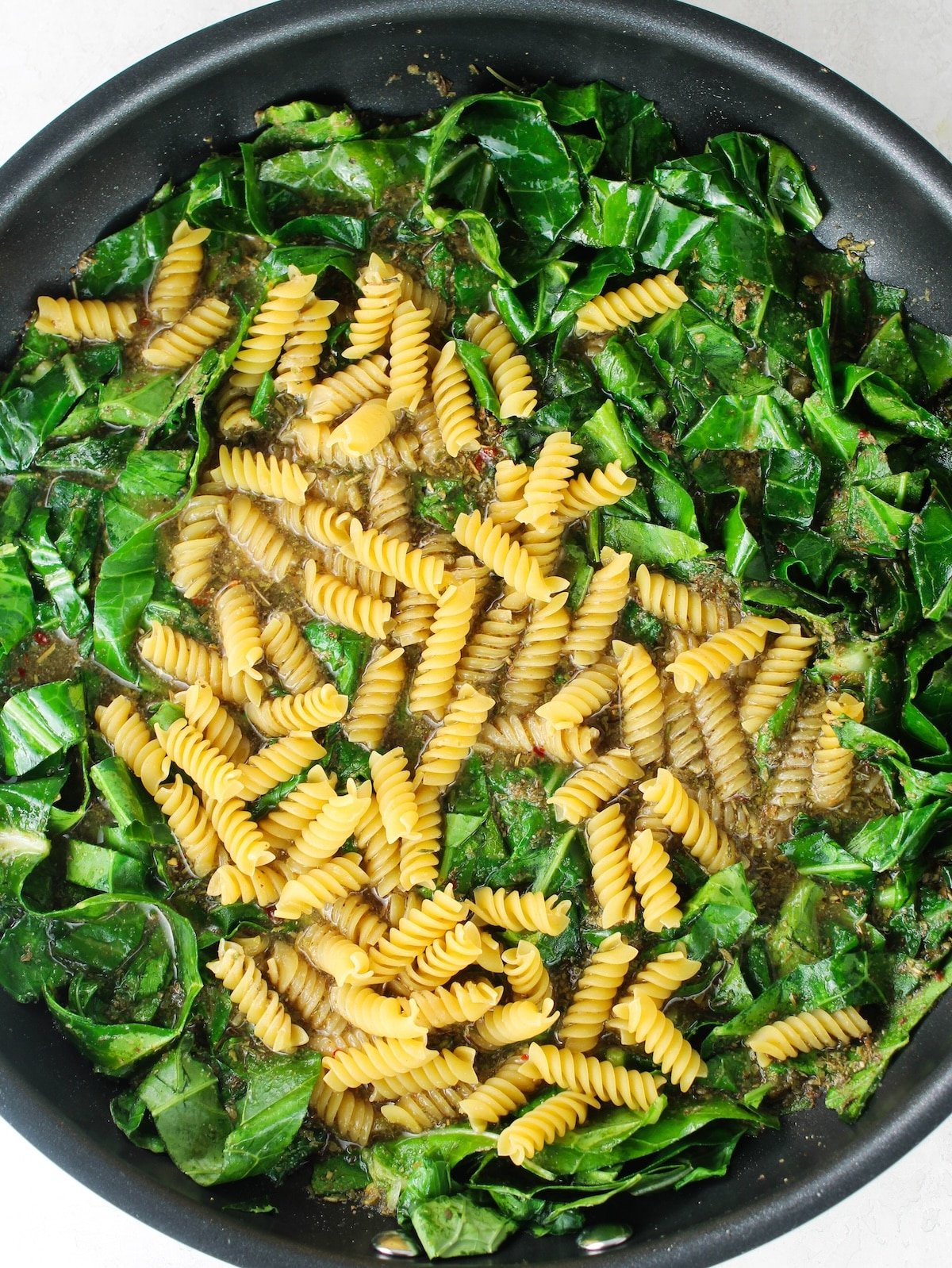 A pan with liquid, seasonings, pasta and greens cooking on the stovetop.