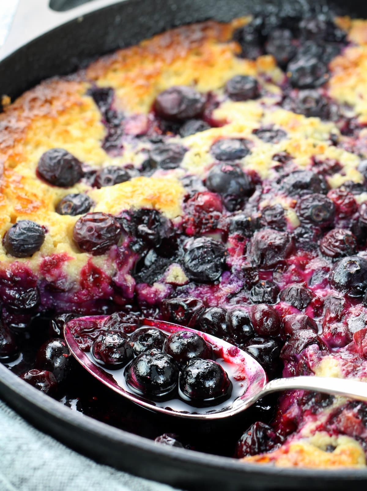 A skillet with baked blueberries and batter with a piece removed.