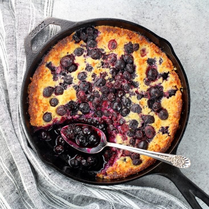 A skillet with baked blueberry cobbler with a piece removed and a spoon holding blueberries.