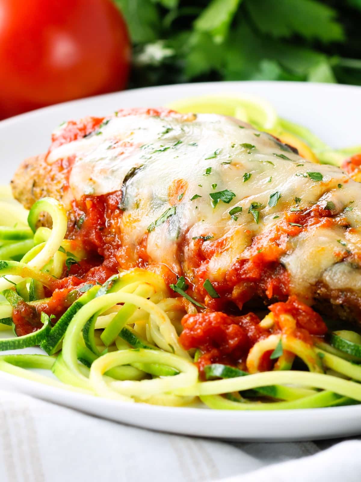 Plate of Low Carb Baked Chicken Parmesan on zucchini noodles A close-up photo of delicious Baked Chicken Parm on a large platter with zoodles, marinara sauce, melted mozzarella cheese, and fresh basil and parsley.