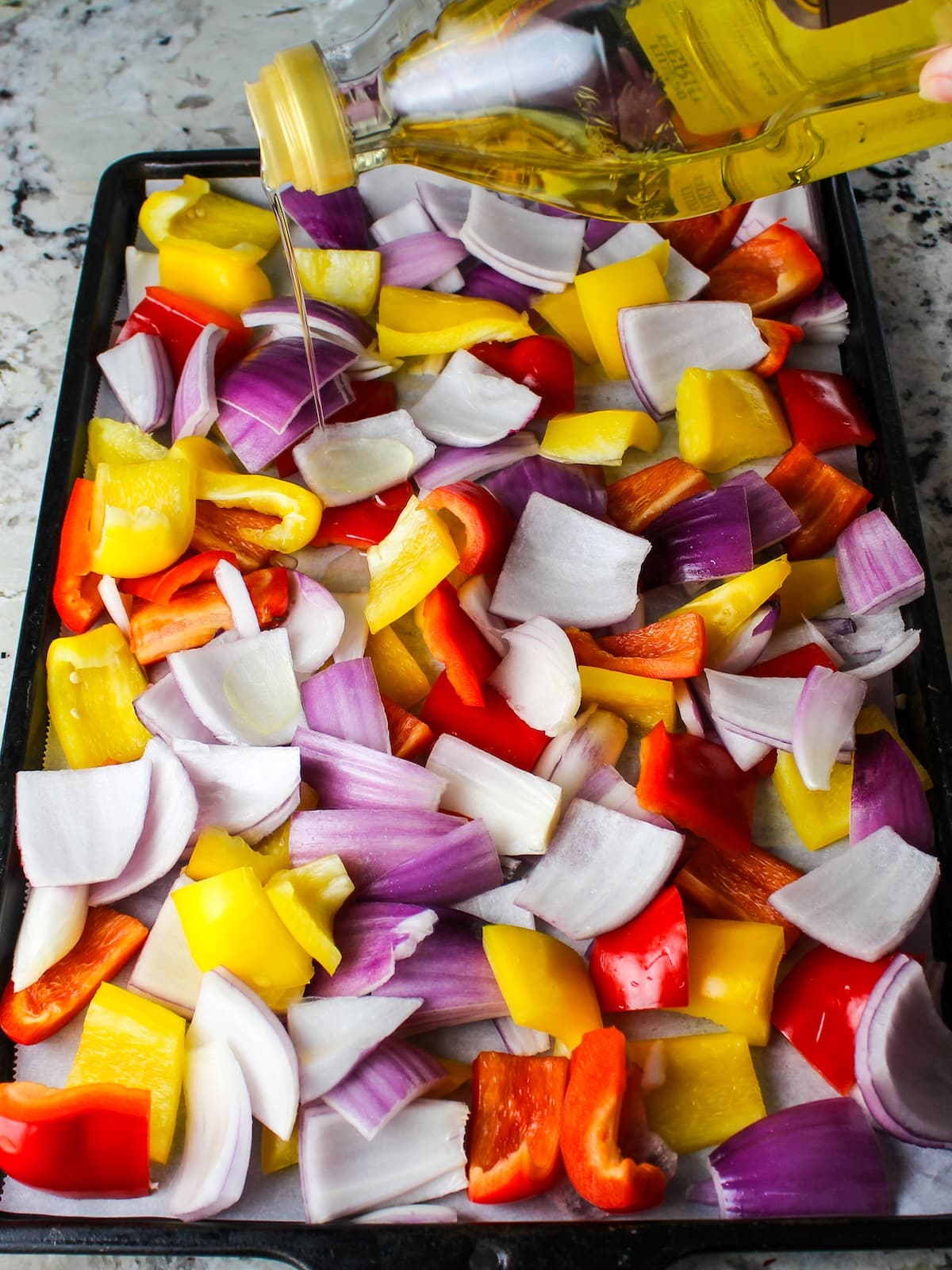 Pouring olive oil over peppers and onions on a sheet pan.