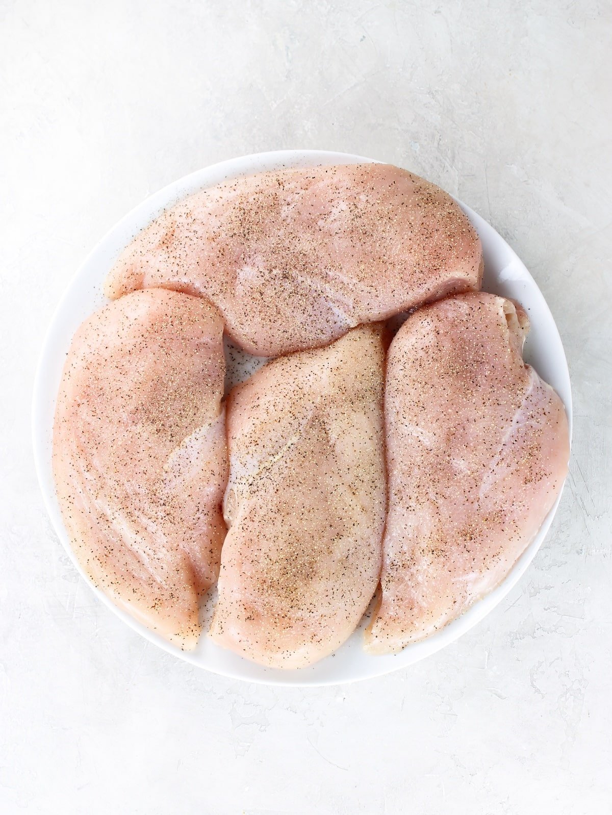 A plate with raw boneless skinless chicken breasts that have been seasoned with salt, pepper and garlic powder.