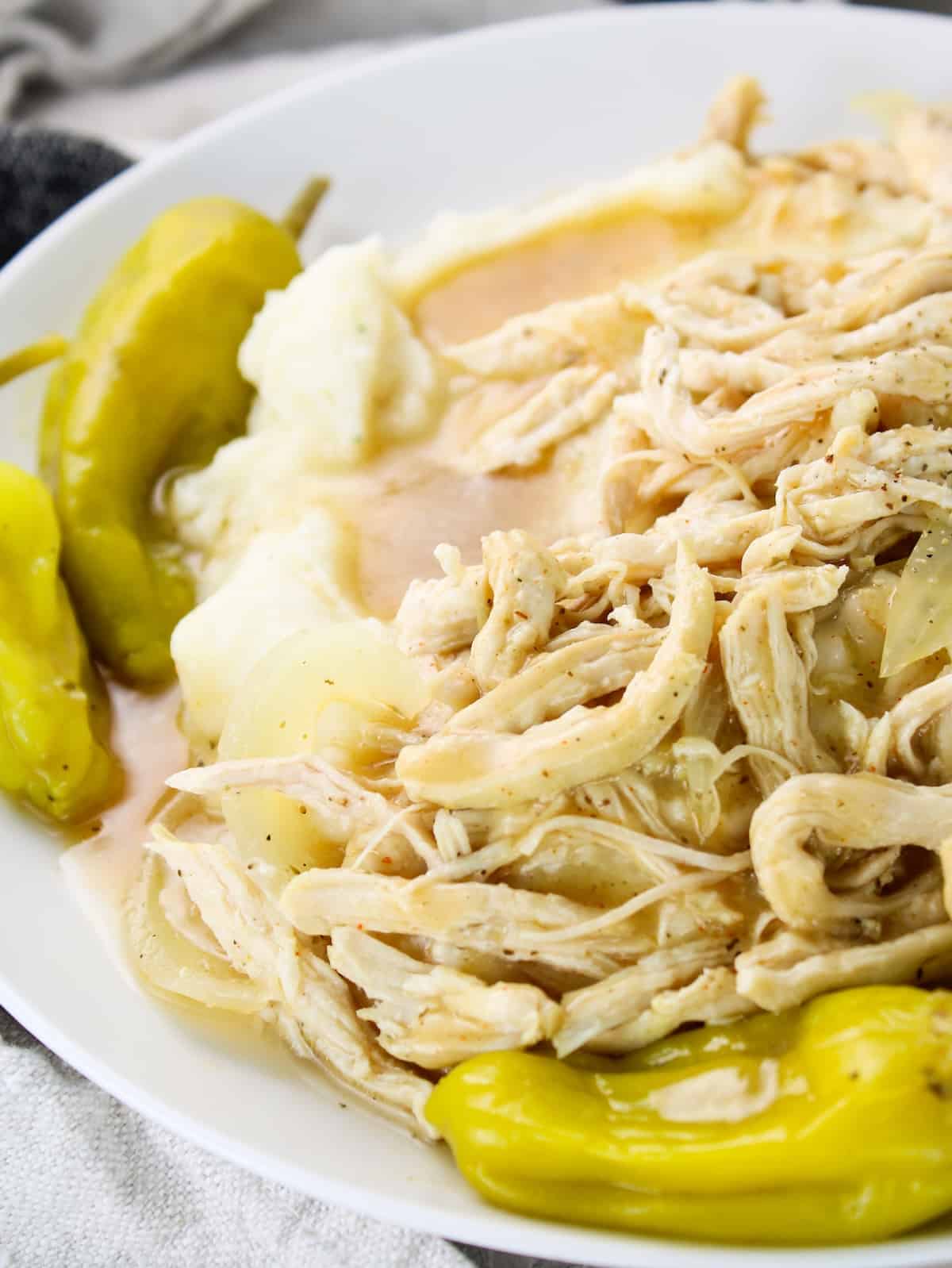 A close-up photo of a plate with Mississippi Chicken, pepperoncini peppers, and cooking liquid served over mashed potatoes.