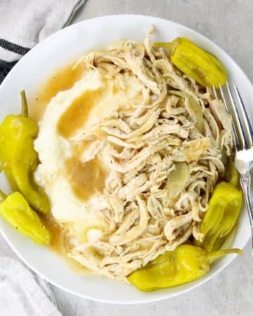 A plate with Mississippi Chicken, pepperoncini peppers, and cooking liquid served over mashed potatoes.
