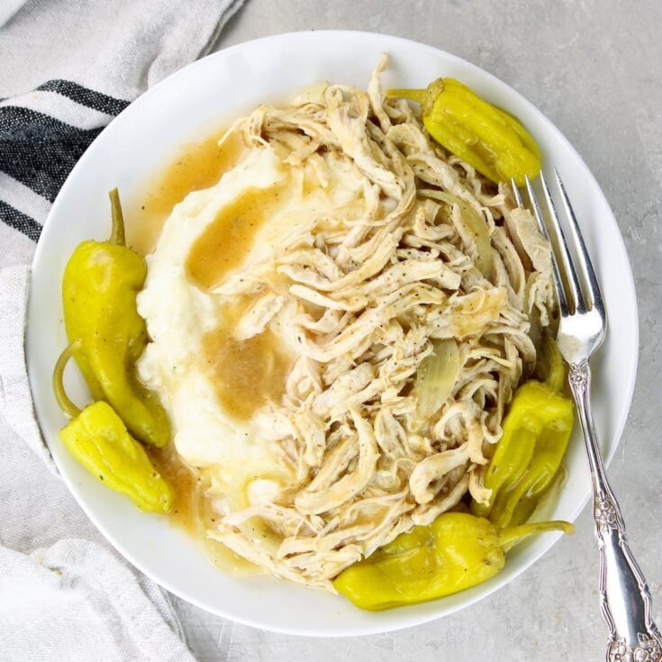 A plate with Mississippi Chicken, pepperoncini peppers, and cooking liquid served over mashed potatoes.