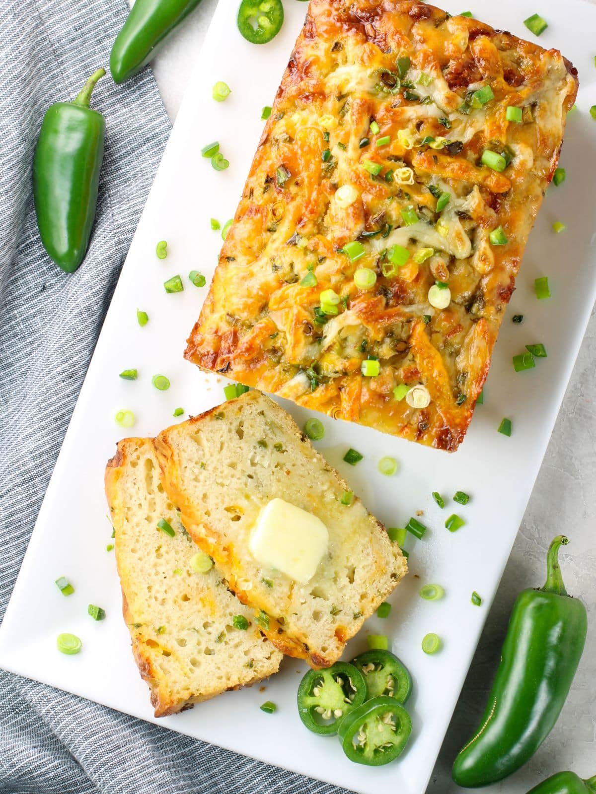 Cheesy jalapeño beer bread loaf on a white platter with a buttered slice cut and fresh jalapeños around it — an easy no-yeast BBQ bread